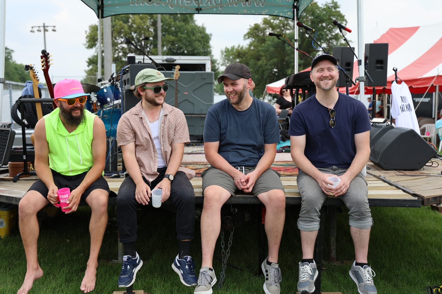 The four members of Bad Friends sitting on the edge of a trailer stage on the grass, laughing between sets at a daytime RAGBRAI show