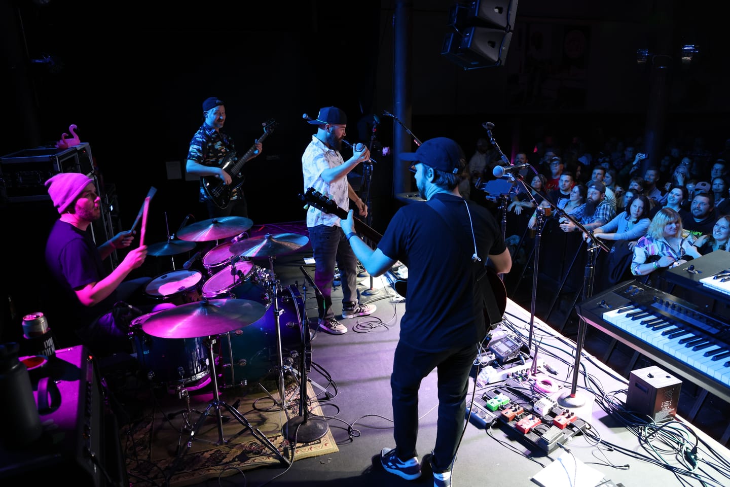 Side-stage view of Bad Friends performing at Wooly's, the drummer lit in pink and the crowd pressed against the barrier